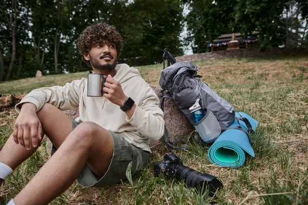 Uomo indiano che tiene thermos tazza e seduto vicino alla fotocamera e allo zaino con attrezzatura da viaggio, premuroso — Foto stock