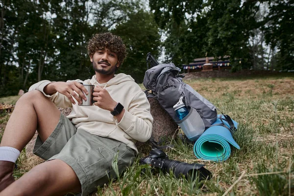 Індійський чоловік тримає термос кухоль і сидить біля камери і рюкзак з туристичним спорядженням, турист — стокове фото