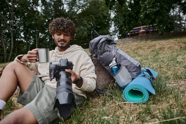 Homem indiano feliz segurando caneca térmica e olhando para fotos na câmera, turista perto de equipamento de viagem — Fotografia de Stock