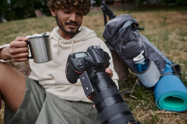 Homem indiano feliz segurando caneca térmica e olhando para fotos na câmera, fotógrafo perto de equipamento de viagem — Fotografia de Stock
