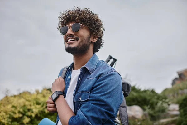 Cheerful indian man in sunglasses standing with backpack on green hill, hiker with travel gear — Stock Photo