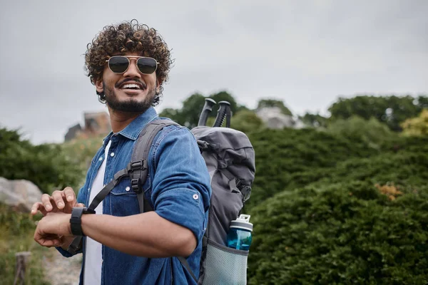 Hombre indio alegre en gafas de sol y camisa de mezclilla comprobar el tiempo en reloj de pulsera, excursionista con equipo de viaje - foto de stock