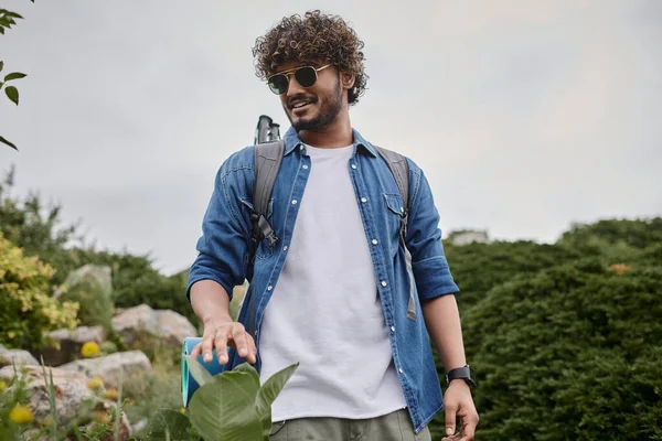 Naturaleza salvaje, hombre indio feliz en gafas de sol caminando con mochila en la colina, excursionista teniendo viaje - foto de stock