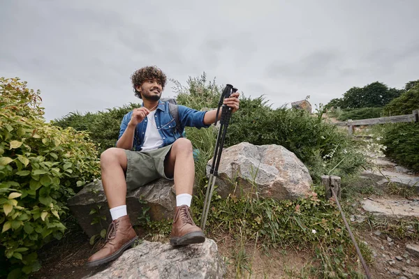 Heureux homme indien assis avec sac à dos sur le rocher et tenant des bâtons de randonnée pendant le trekking, aventure — Photo de stock