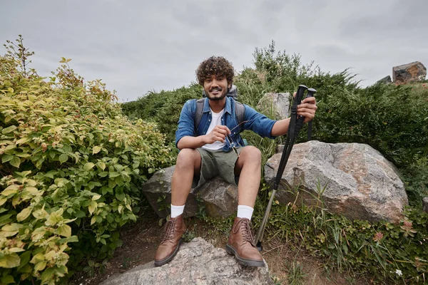 Indiana homem sentado com mochila no rock e segurando caminhadas varas durante trekking, aventura — Fotografia de Stock
