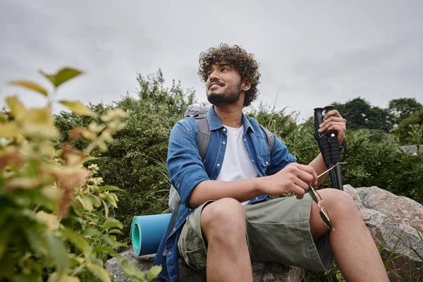 Glücklicher indischer Mann sitzt mit Rucksack und hält Wanderstöcke während des Trekkings, wilde Natur — Stockfoto