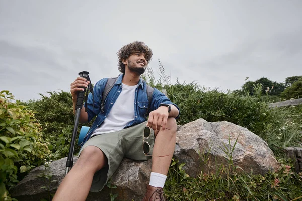 Glücklich indischer Tourist sitzt mit Rucksack und hält Wanderstöcke während des Trekkings, wilde Natur — Stockfoto
