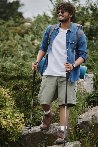 Happy indian hiker in sunglasses holding hiking sticks and walking with backpack during trekking — Stock Photo