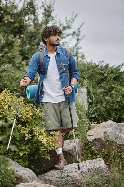Curly indian tourist walking with backpack and holding hiking sticks during trekking, wild nature — Stock Photo