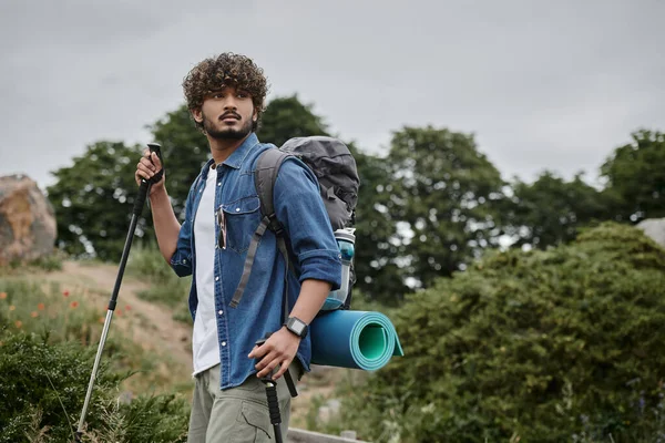 Curly indian backpacker holding trekking poles on path in woods, hiker having adventure concept — Stock Photo
