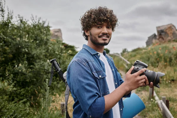 Concepto de viaje y fotografía, mochilero indio rizado que sostiene la cámara digital durante el viaje por la naturaleza - foto de stock