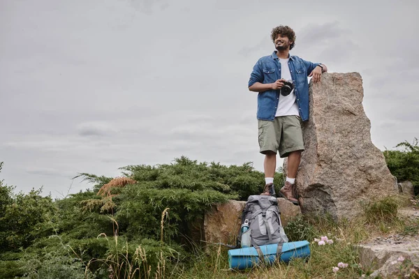 Fotografía y concepto de la naturaleza, mochilero indio feliz sosteniendo cámara digital y de pie sobre roca - foto de stock