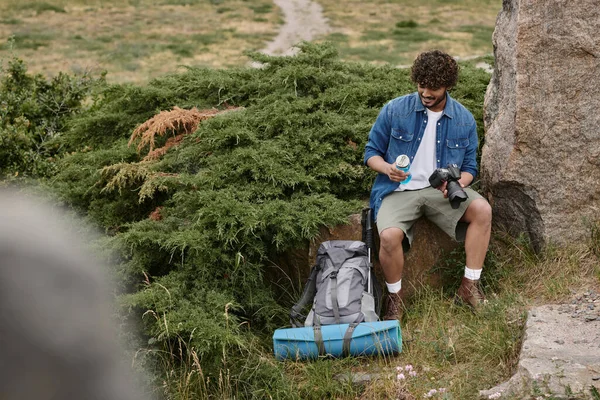 Tourist and nature concept, indian man holding bottle with water and camera while sitting on rock — Stock Photo