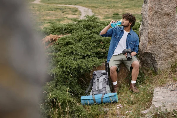 Tourist and nature concept, indian man drinking water and holding camera while sitting on rock — Stock Photo