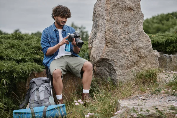 Travel and nature concept, indian man holding bottle and using digital camera while sitting on rock — Stock Photo