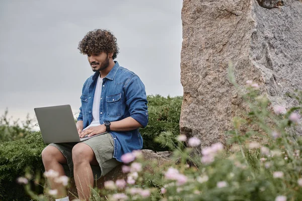 Digital nomad concept, curly indian man using laptop and sitting on rock in natural place — Stock Photo