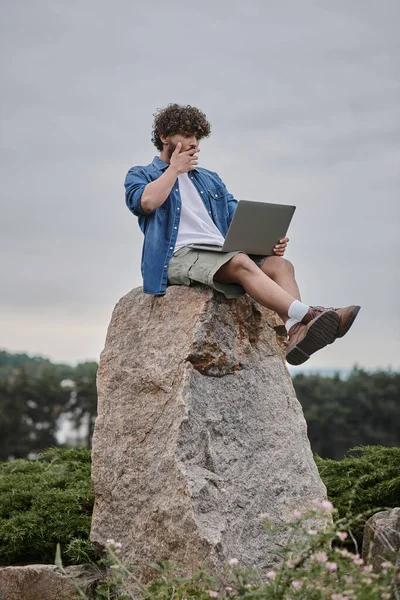 Digital nomad concept, shocked indian freelancer covering mouth and using laptop, sitting on rock — Stock Photo