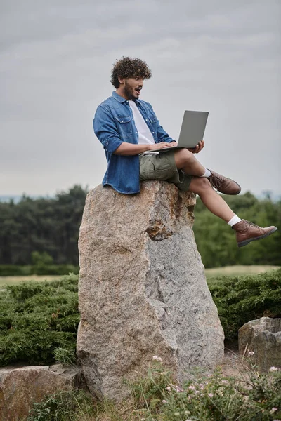 Digital nomadism, amazed indian freelancer using laptop, sitting on rock, remote work concept — Stock Photo