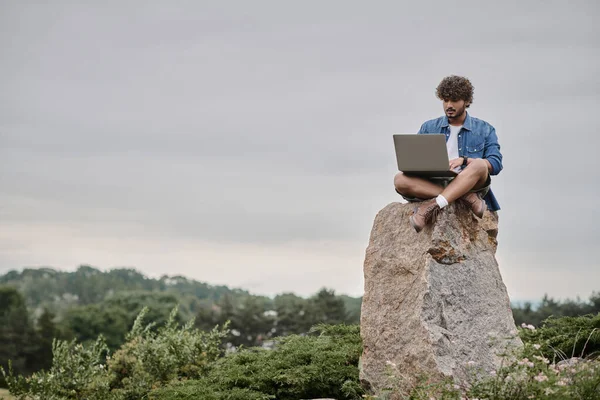 Nómada digital, hombre indio rizado usando el ordenador portátil, sentado en la roca, concepto de trabajo remoto, vista del paisaje - foto de stock