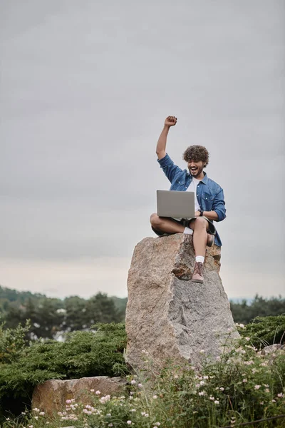 Концепція цифрової кочівниці, збуджений індійський чоловік святкує перемогу під час використання ноутбука, сидячи на скелі — стокове фото