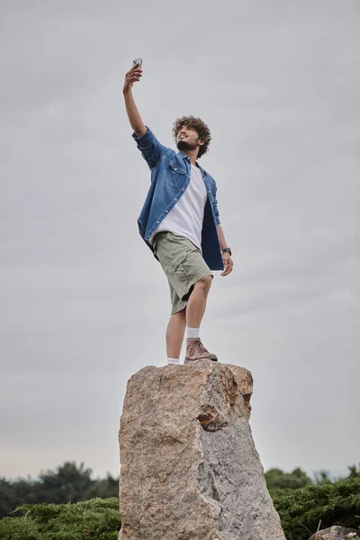 Nomadism concept, curly indian man standing on rock and holding smartphone, searching signal — Stock Photo