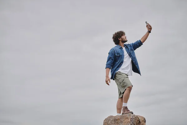 Nomadentum-Konzept, lockiger Indianer, der auf einem Felsen steht und Signal sucht, während er sein Smartphone in der Hand hält — Stockfoto