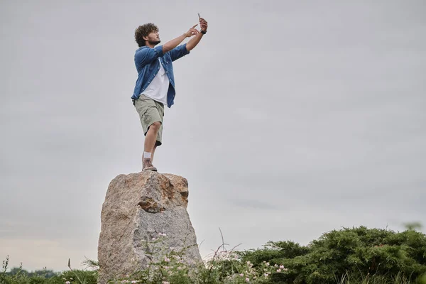 Concepto de nomadismo, el hombre indio rizado de pie en el rock y la señal de búsqueda mientras sostiene el teléfono móvil - foto de stock