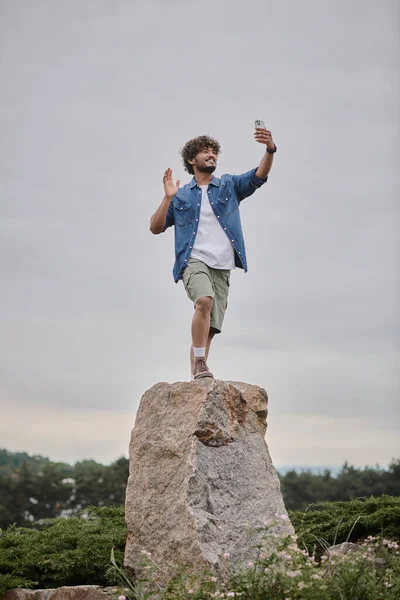Концепція кочовиділення, кучерявий індійський чоловік, що стоїть на камені і має відеочат на смартфоні, хвильова рука — стокове фото