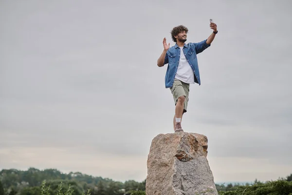 Nomadism concept, happy indian man standing on rock and having video chat on smartphone, wave hand — Stock Photo