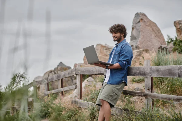 Concepto nómada digital, hombre indio alegre de pie cerca de valla de madera y el uso de ordenador portátil, la naturaleza - foto de stock