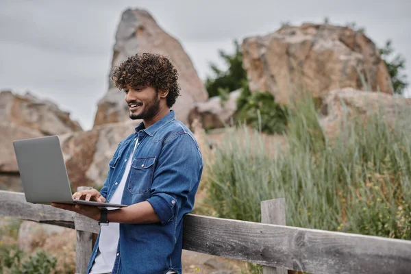 Digitales Nomadenkonzept, fröhlicher Indianer, der am Holzzaun steht und Laptop benutzt, Landschaft — Stockfoto