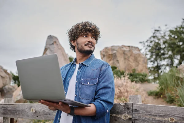 Концепція цифрової кочівниці, позитивний індійський чоловік стоїть біля дерев'яного паркану і використовує ноутбук, сільську місцевість — стокове фото