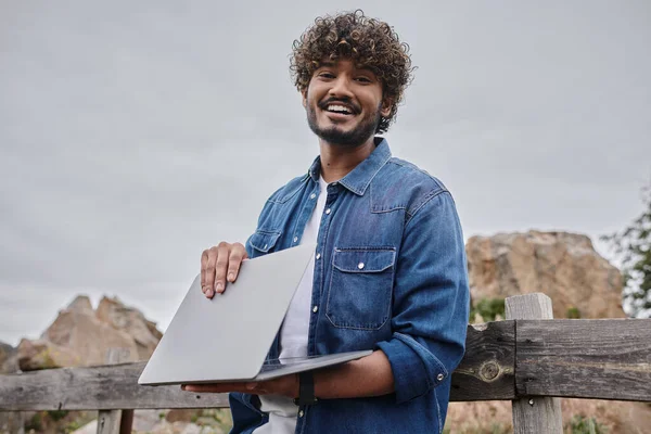 Fröhlicher indischer Mann, der in der Nähe eines Holzzaunes in der Landschaft steht und Laptop, digitaler Nomade benutzt — Stockfoto