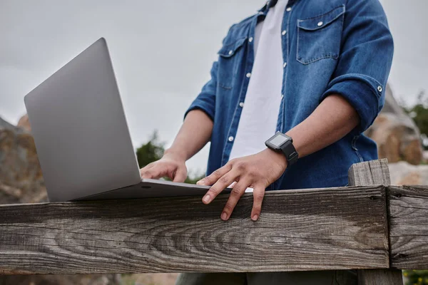 Обрезанный вид фрилансера, стоящего рядом с деревянным забором и использующего ноутбук, цифровую концепцию кочевника — стоковое фото