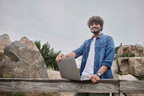 Hombre indio positivo de pie cerca de valla de madera en el campo y el uso de ordenador portátil, nomadismo digital - foto de stock