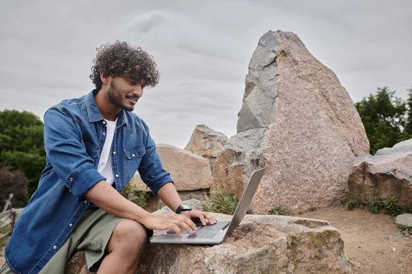 Hombre indio feliz escribiendo en el ordenador portátil y trabajando remotamente en lugar natural, nomadismo digital - foto de stock
