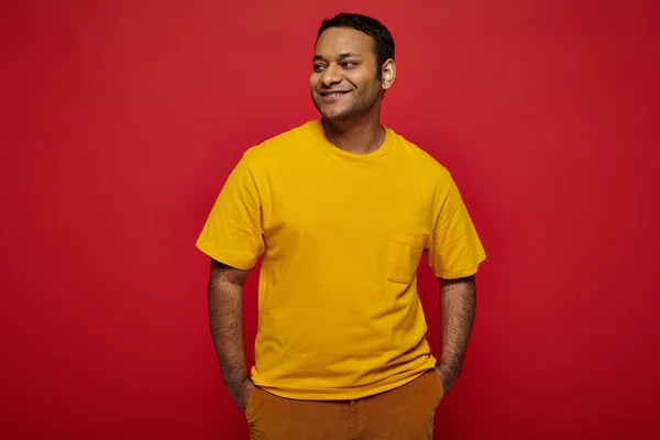 Позитивний індійський чоловік у яскравому повсякденному вбранні, що стоїть і посміхається на червоному тлі в студії — стокове фото