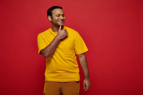 Hombre indio feliz en ropa casual brillante de pie y sonriendo sobre fondo rojo en el estudio - foto de stock