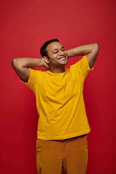 Homem indiano satisfeito em roupas casuais brilhantes de pé e sorrindo no fundo vermelho, postura relaxada — Fotografia de Stock