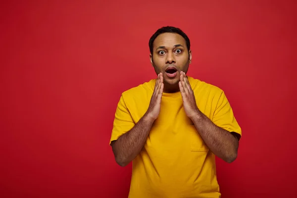 Shocked indian man in bright casual clothes looking at camera on red background, open mouth — Stock Photo