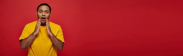 Homem indiano chocado em roupas casuais brilhantes olhando para a câmera no fundo vermelho, boca aberta, banner — Fotografia de Stock
