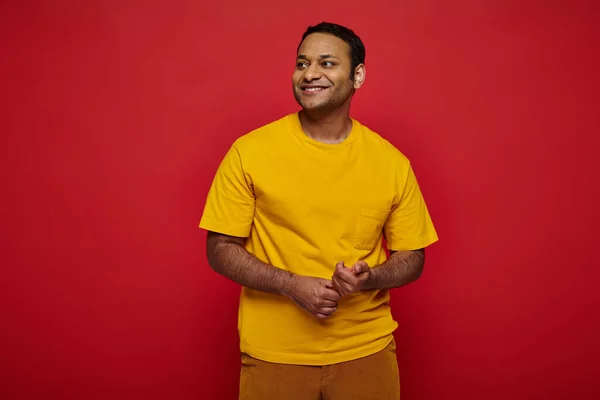 Homem indiano delicioso em roupas casuais brilhantes olhando para longe e sorrindo no fundo vermelho no estúdio — Fotografia de Stock