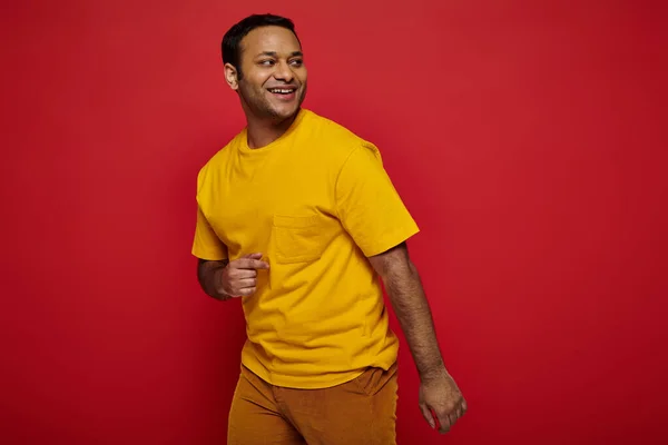 Homem indiano alegre em roupas casuais brilhantes olhando para longe e sorrindo no fundo vermelho, otimista — Fotografia de Stock