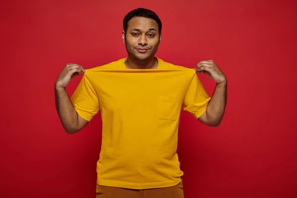 Homme indien à la mode touchant t-shirt jaune et regardant la caméra sur fond rouge, style personnel — Photo de stock