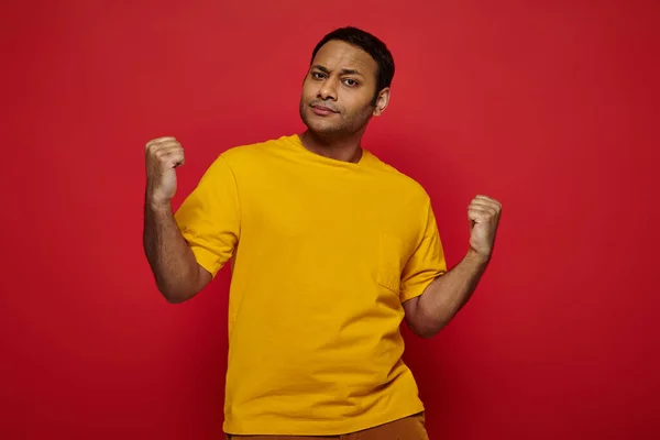 Expressive indian man in casual clothes standing with clenched fists  on red background, strength — Stock Photo