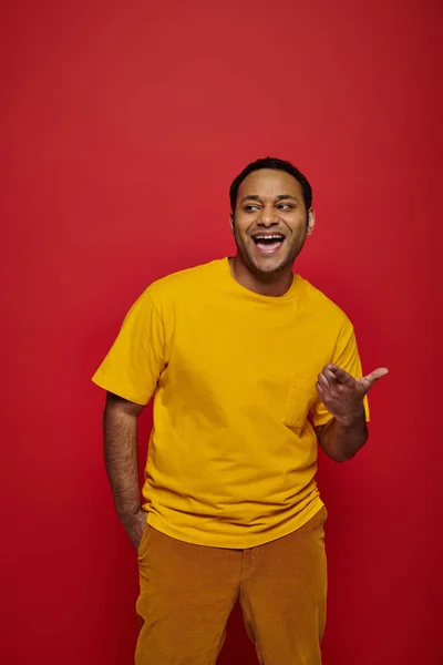 Smiling indian man in bright casual clothes mocking and pointing with finger on red background — Stock Photo