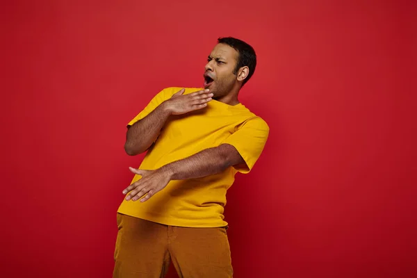 Hombre indio emocional en ropa casual brillante que muestra una reacción expresiva sobre el fondo rojo, wow - foto de stock