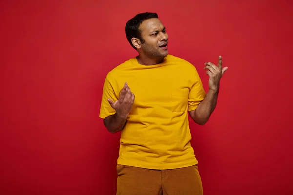 Homme indien émotionnel en vêtements décontractés lumineux gestuelle tout en expliquant quelque chose sur fond rouge — Photo de stock