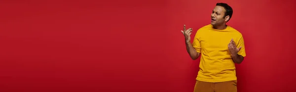 Homme indien émotionnel en vêtements décontractés lumineux gestuelle tout en expliquant quelque chose sur rouge, bannière — Photo de stock