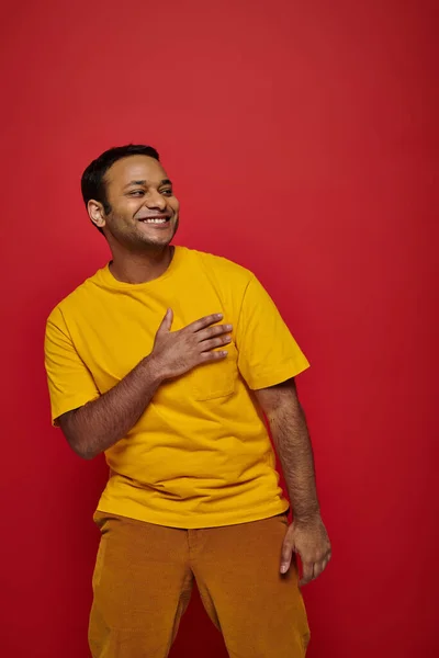 Благодарный индийский мужчина в яркой повседневной одежде улыбается и смотрит в сторону на красном фоне в студии — Stock Photo